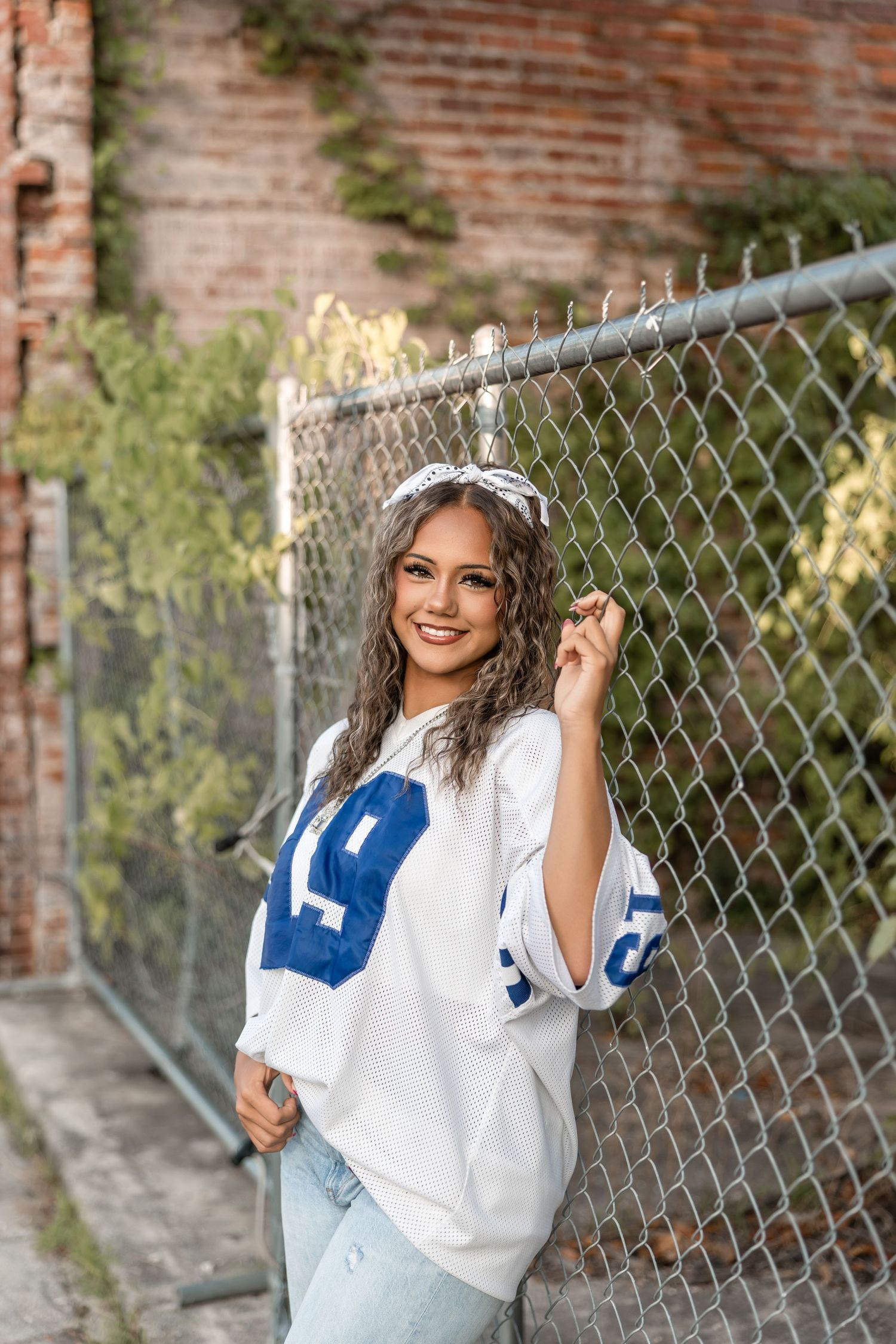 A person wearing a white and blue varsity-style sweater poses casually against a chain link fence near a brick wall.
