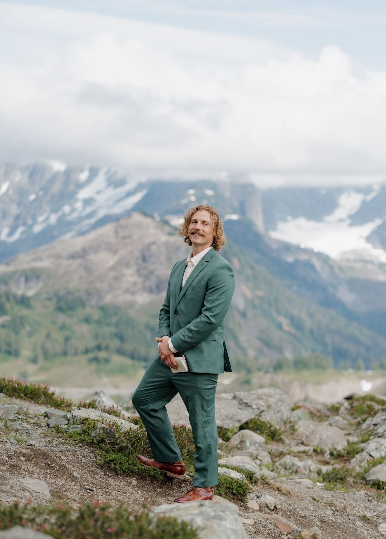 A person in a green suit stands on a rocky mountain ridge with snow-capped peaks in the background.