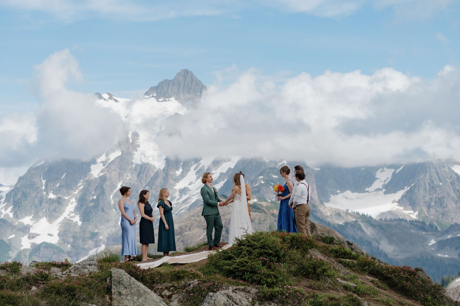 Hikers stand on a mountain ridge overlooking snow-capped peaks and dramatic alpine scenery in the distance.