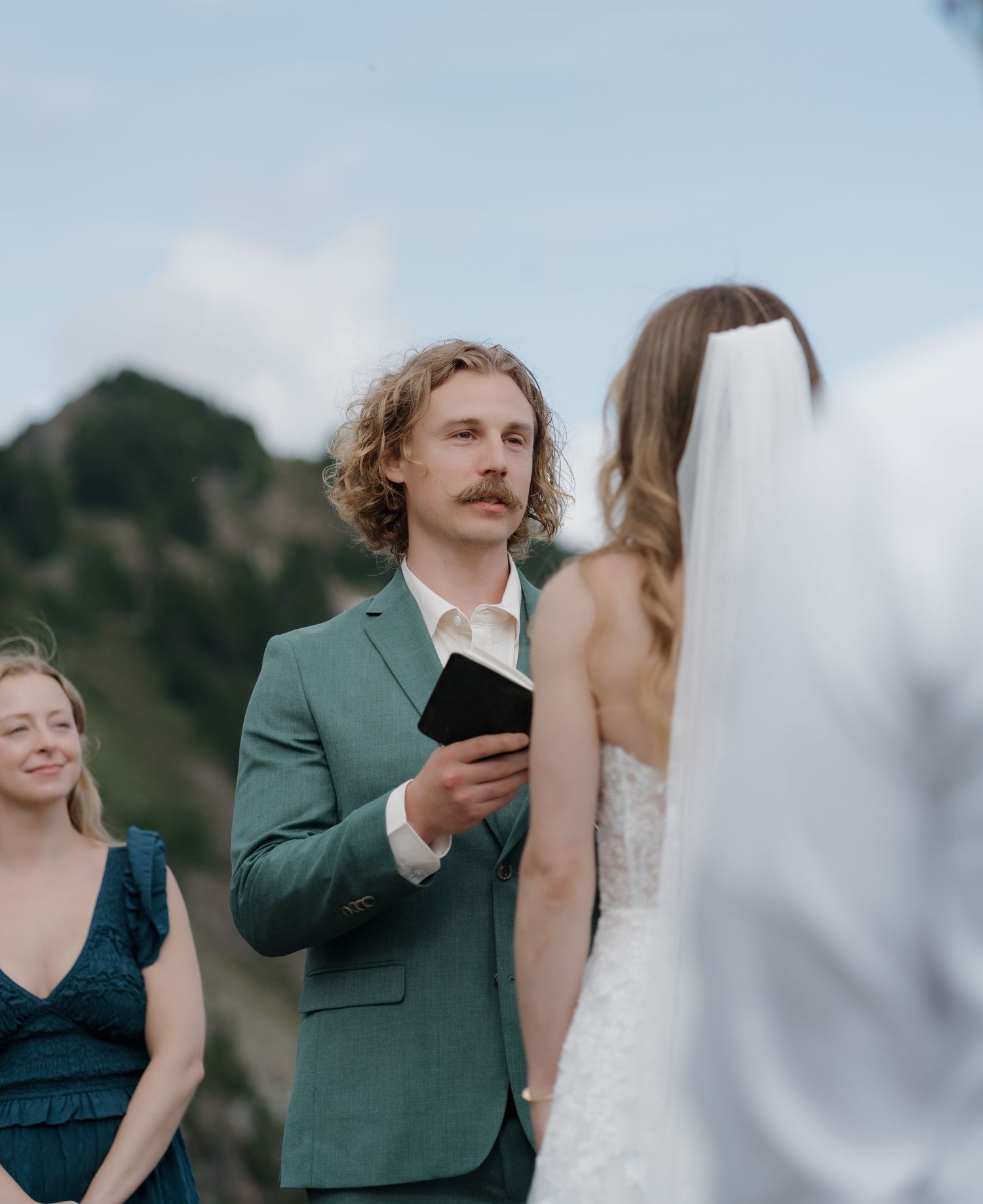 Wedding couple shares intimate moments during outdoor ceremony while guests look on in teal and green attire.
