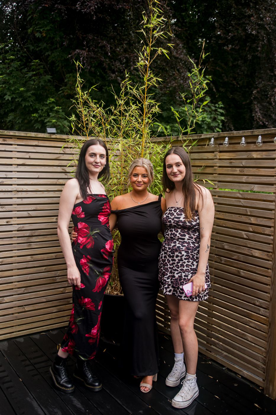 Three guests pose at outdoor party against wooden fence backdrop with bamboo plants.