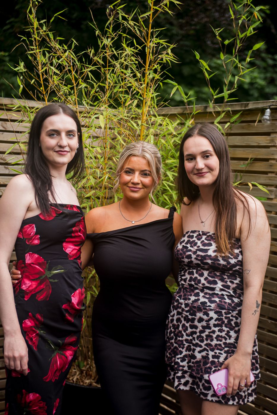 Three party guests pose outdoors against wooden fence with bamboo plants.