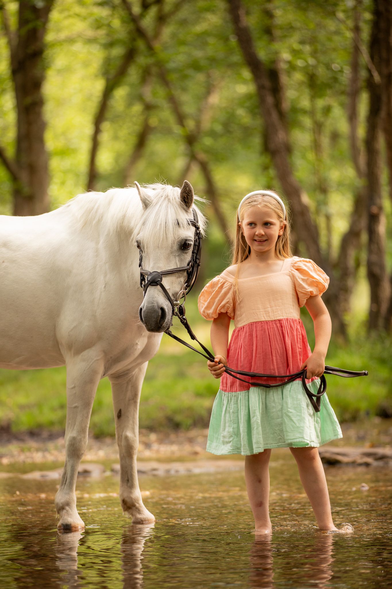 A white pony and someone in colorful clothing stand in shallow water on a sunny day in a wooded area.