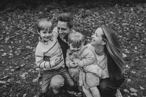 Black and white photo of a family of four enjoying a playful moment together while sitting on fallen autumn leaves.
