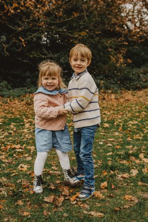 Two children holding hands and smiling while standing on autumn leaves in a park setting.