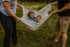 A baby is being gently rocked in a white hammock outdoors while people gather around on a sunny day.
