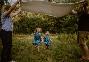 Two children in blue dresses jumping with joy as adults hold up a white sheet in a grassy field at sunset.