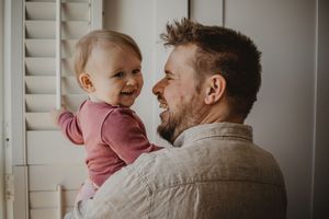 Parent and baby share a tender moment near white shuttered windows in natural light.