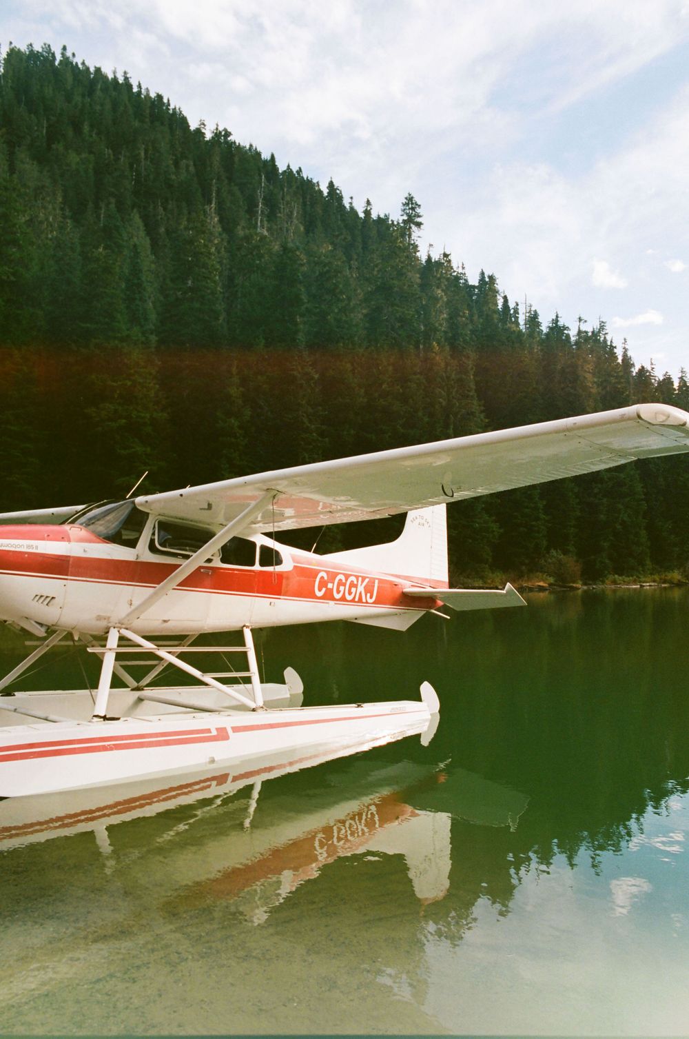 Red and white floatplane docked on calm mountain lake with evergreen forest backdrop.