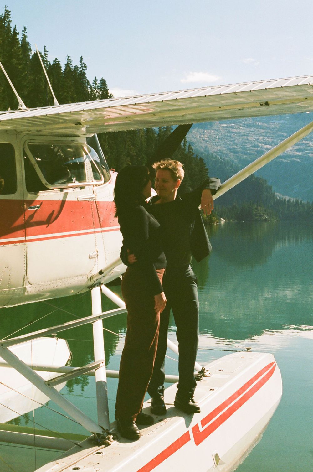 Figure in dark clothing poses on the pontoon of a red and white seaplane on a scenic mountain lake.