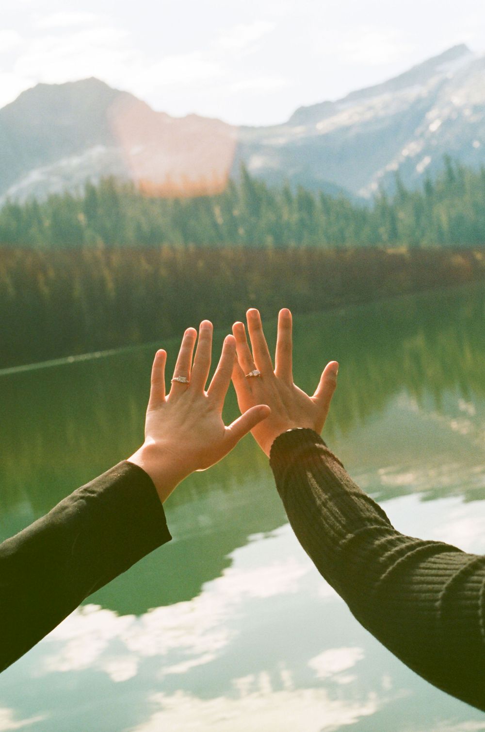 Hands reach towards a serene mountain lake at sunset with evergreen forest reflections in the calm waters.