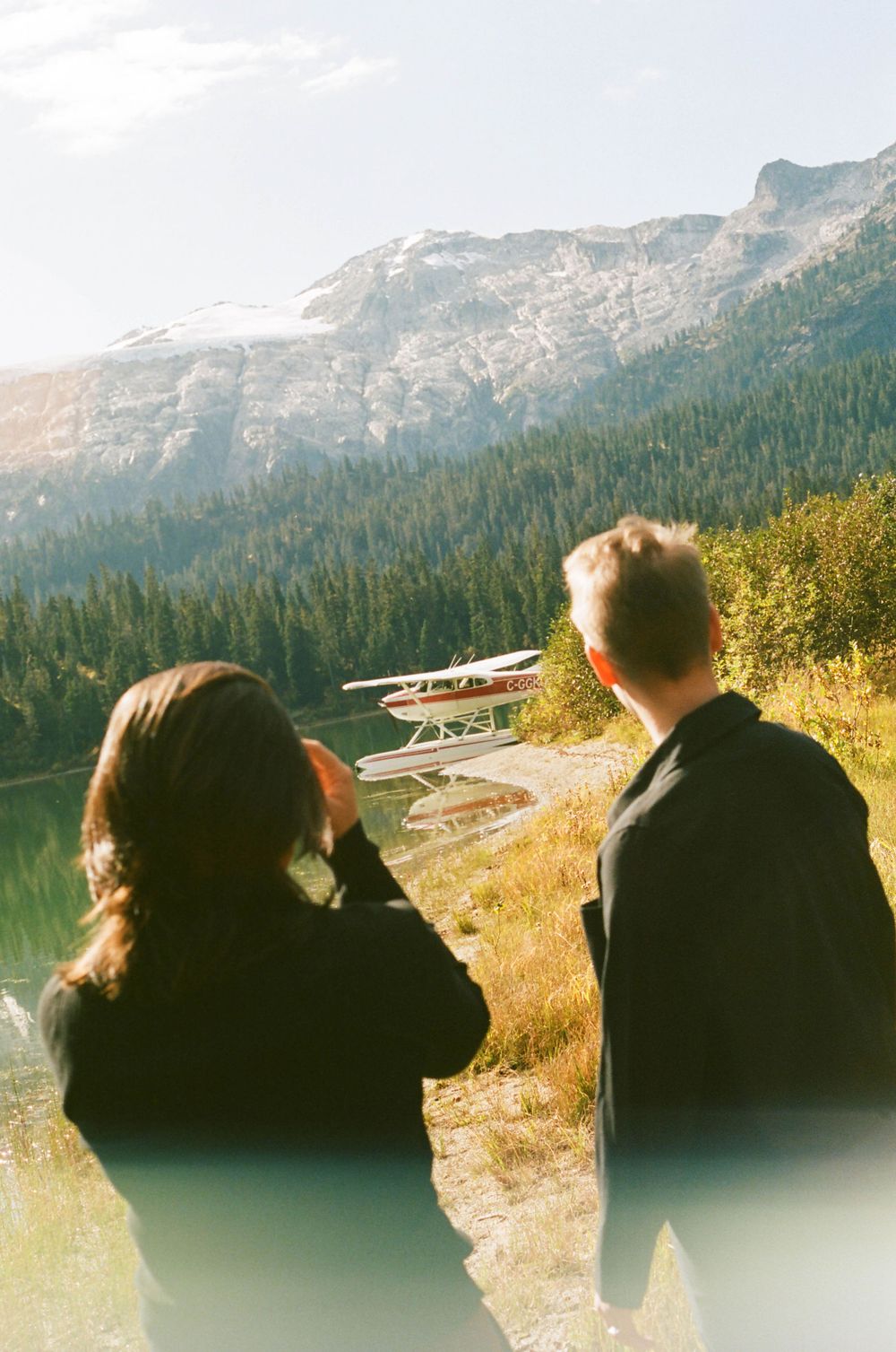 Two people admiring a scenic mountain lake view with a seaplane docked along the shoreline in the distance.