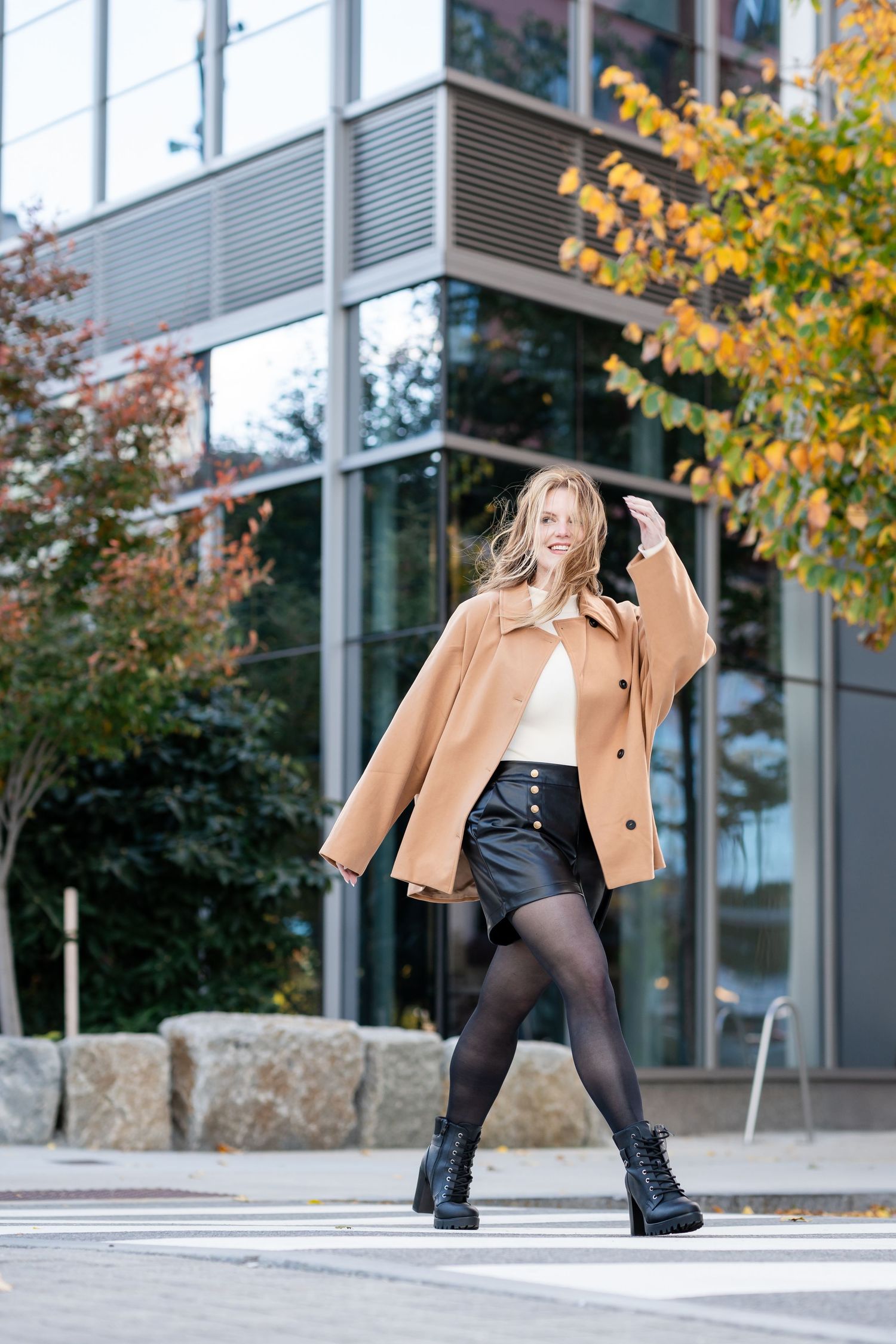 A stylish fall fashion photoshoot featuring a beige coat and black boots against an urban backdrop with autumn foliage.