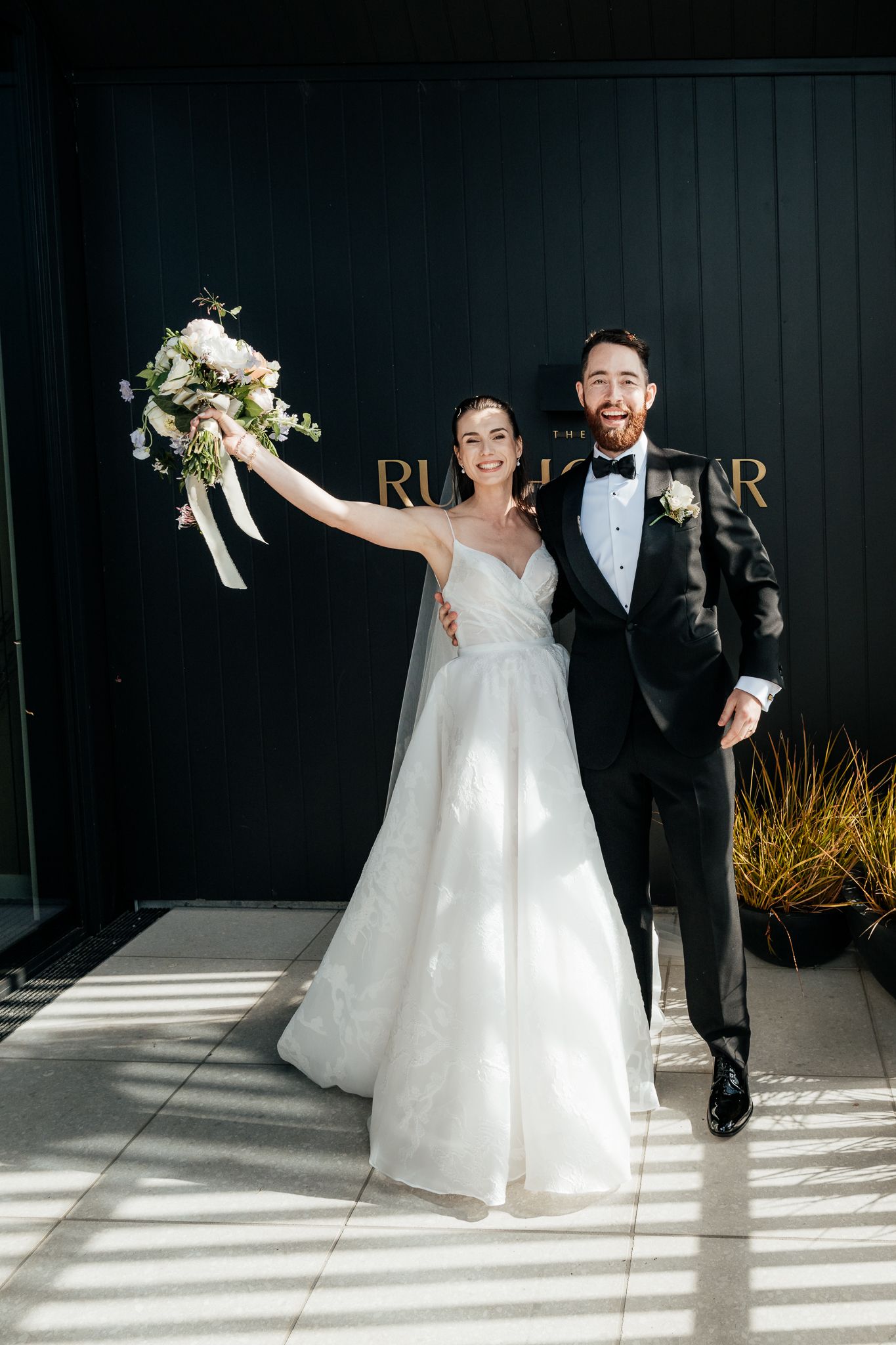 A joyful wedding couple pose together outside the Runholder, with the bride holding up a white floral bouquet by August and Vine