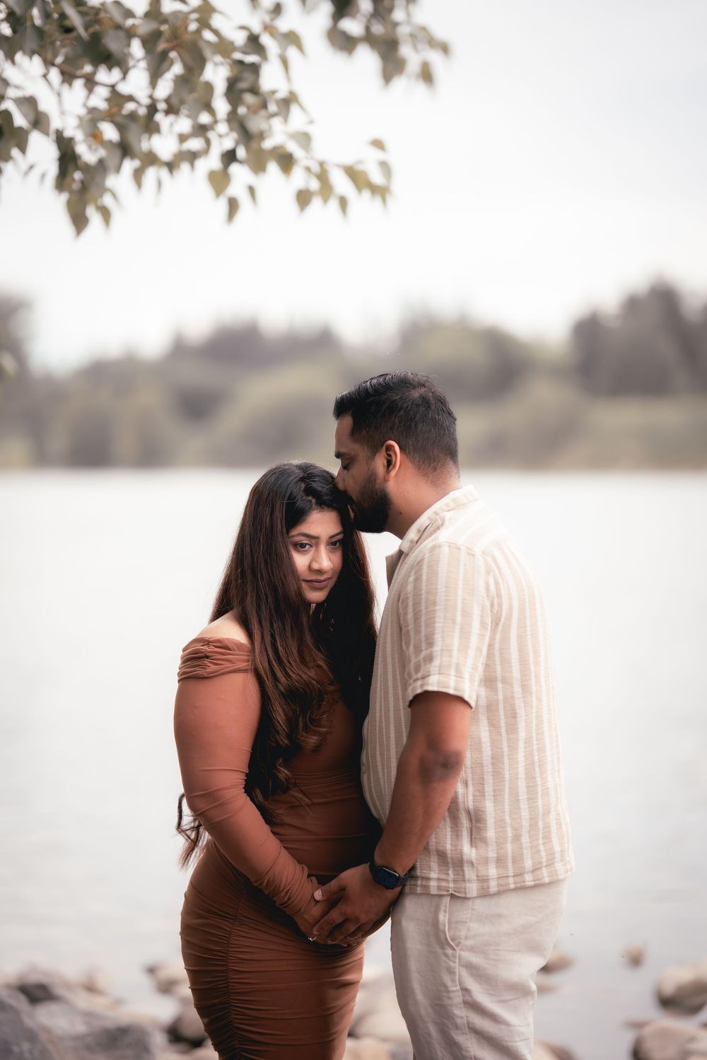 A couple shares an intimate moment by a serene lakeside during a maternity photoshoot in soft natural lighting.