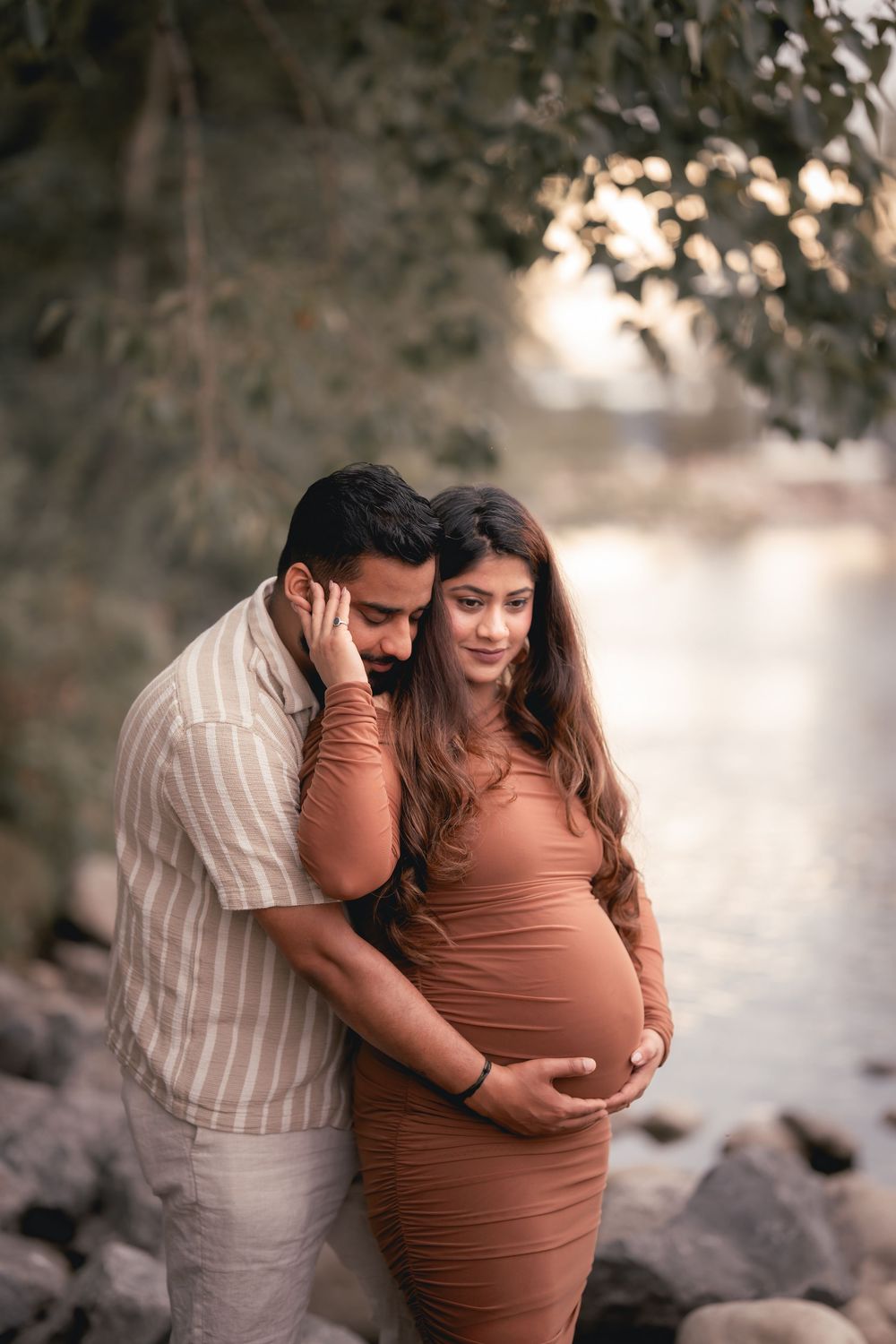 Couple embraces tenderly during maternity photoshoot outdoors in natural lighting with brown and neutral tones.