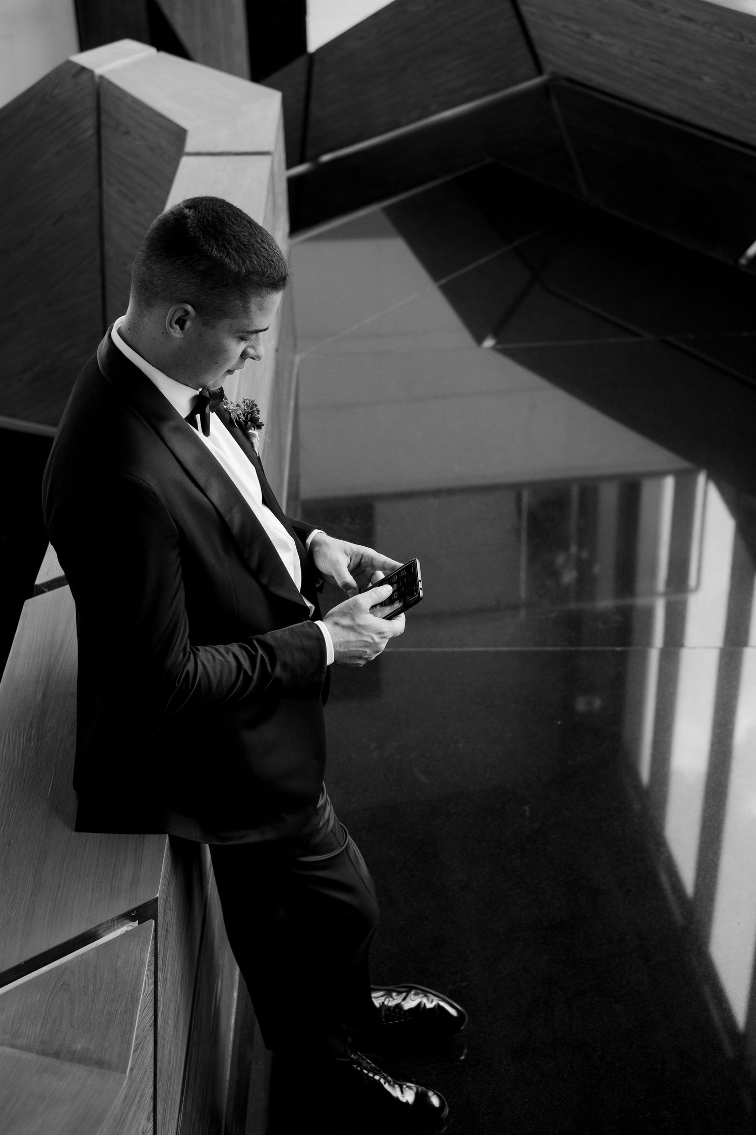 Black and white photo of person in formal tuxedo checking phone while standing in modern architectural setting.