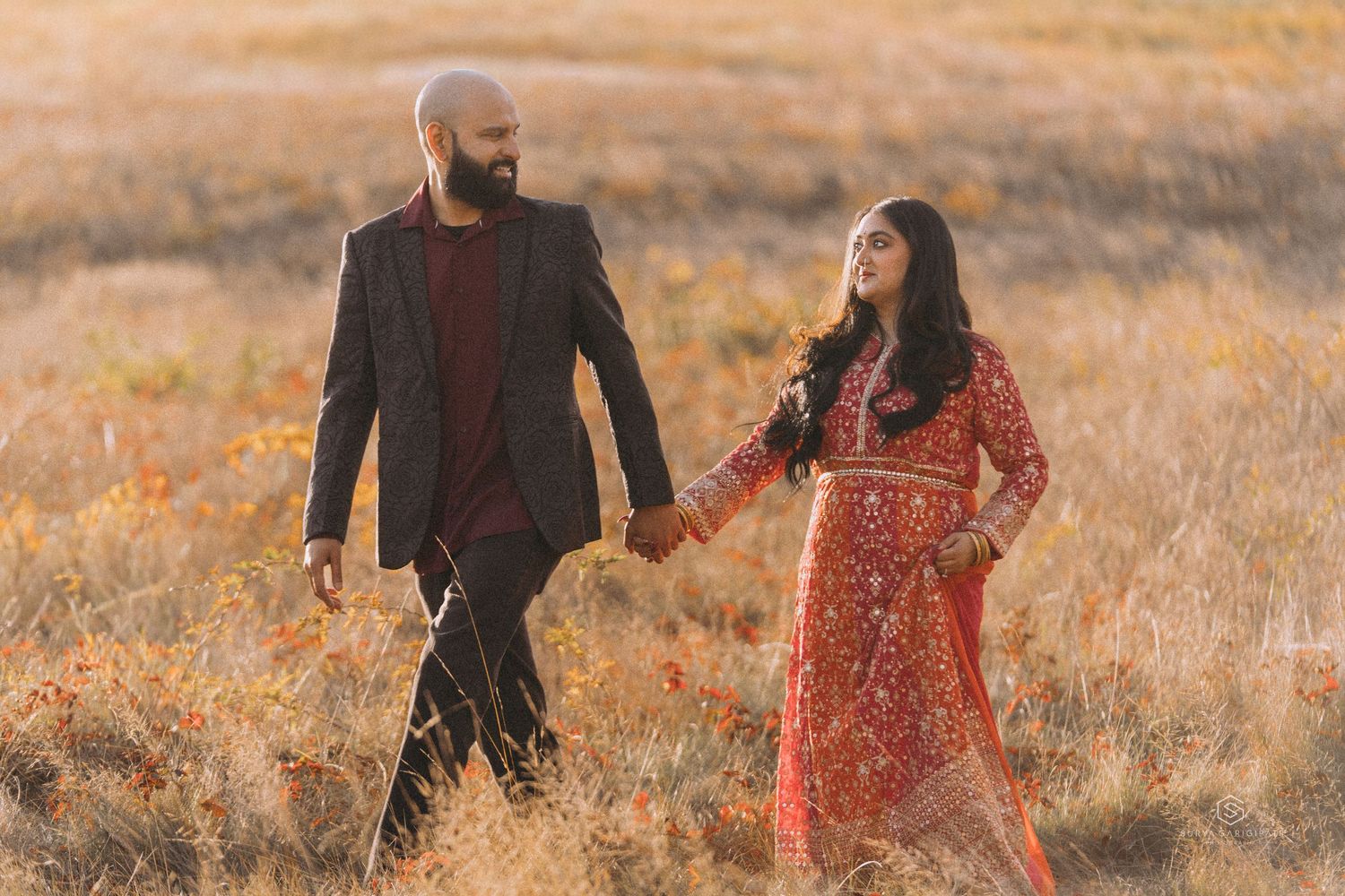 Couple walking together in a golden autumn field wearing burgundy and floral outfits during sunset.