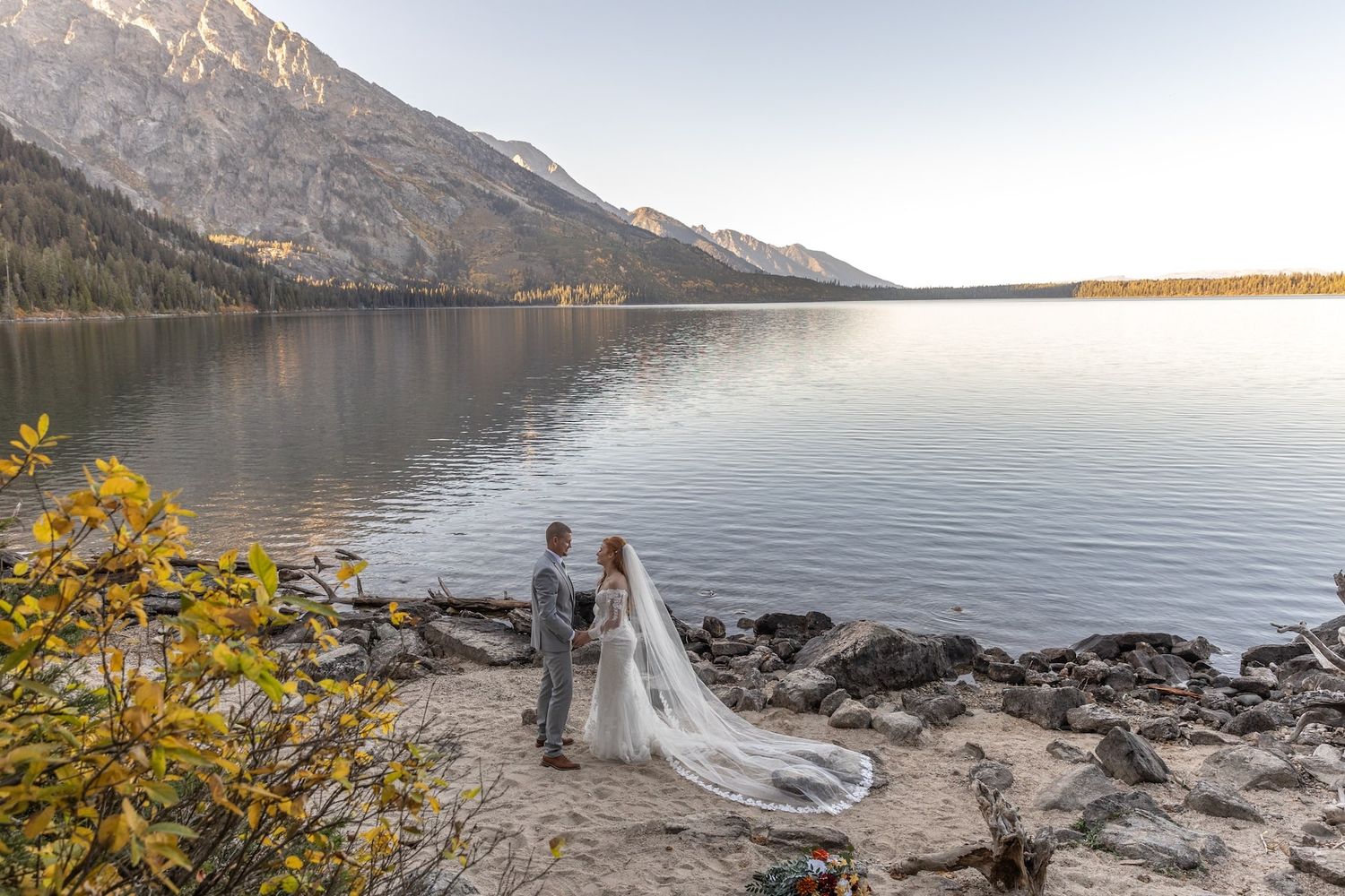 A couple shares a romantic moment by a river with golden autumn trees and majestic mountains in the background.