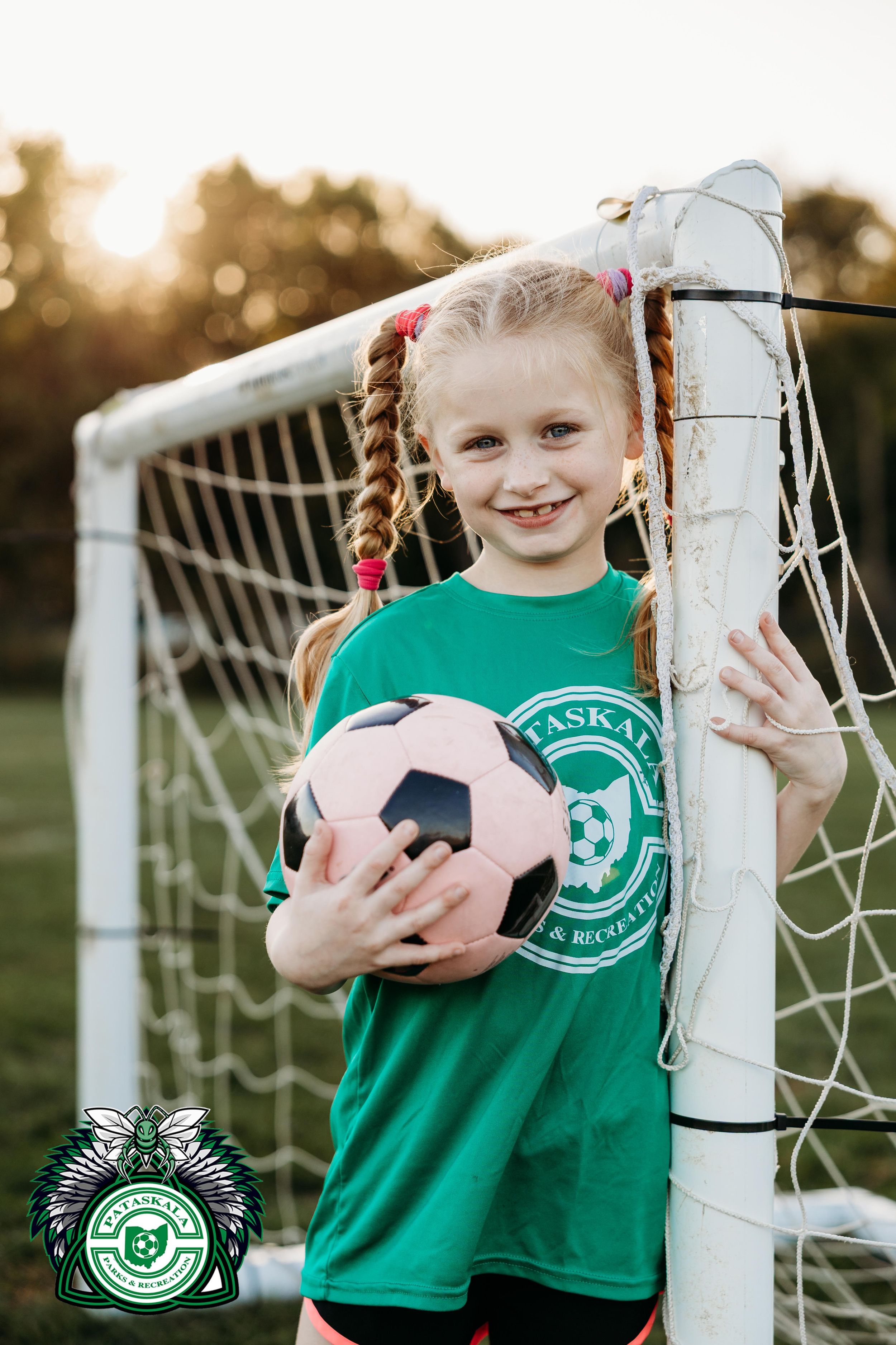 Young soccer player poses with ball next to goal post at sunset wearing green jersey.