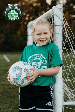 Young soccer player in green jersey poses with soccer ball by goal post in outdoor setting.