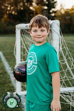 Young soccer player in green uniform holds a ball while standing next to a goal post on a grass field at sunset.