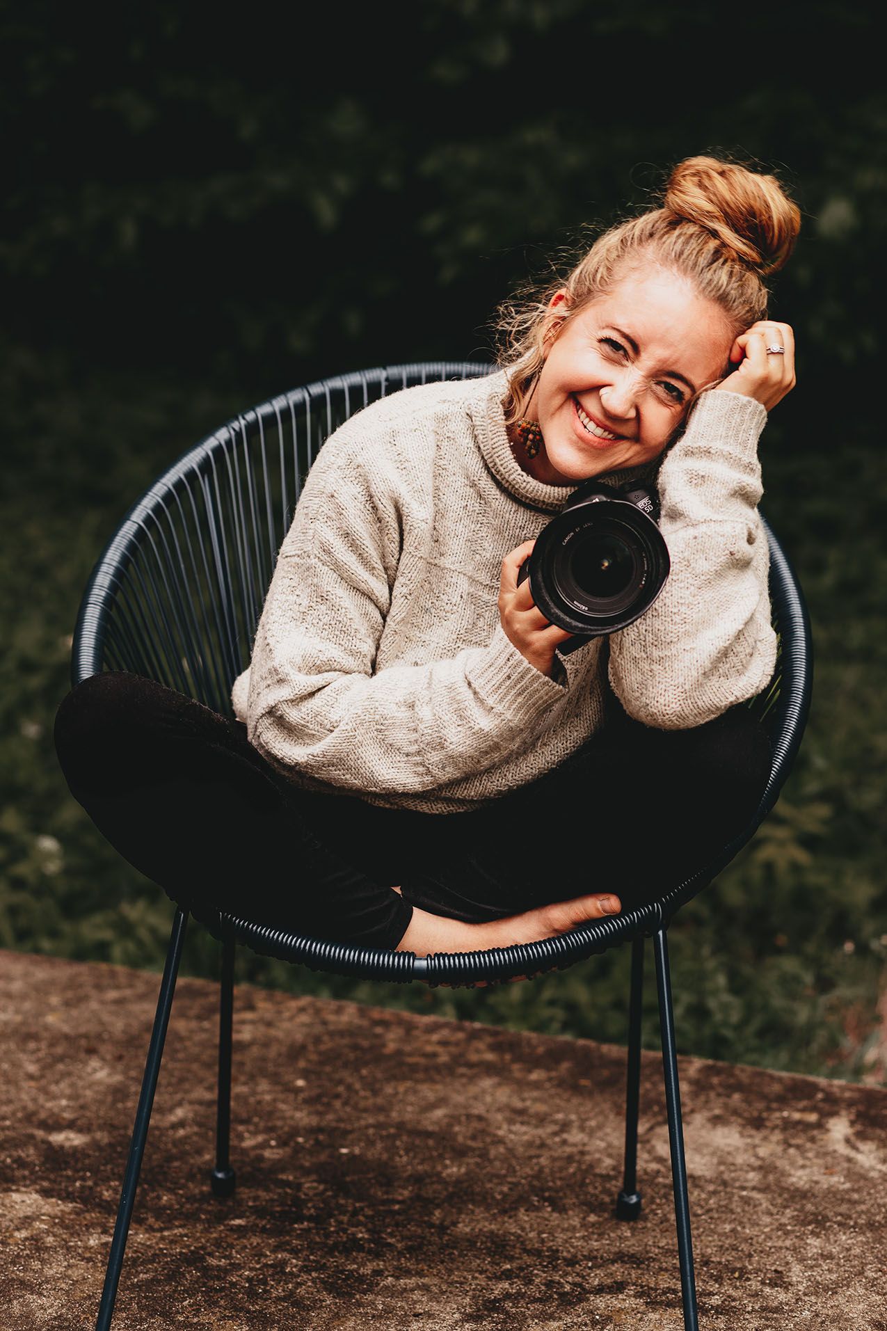 Miranda, a white woman, sits cross-legged in a chair smiling. She holds a camera in her hand and is a Twin Cities family photographer.