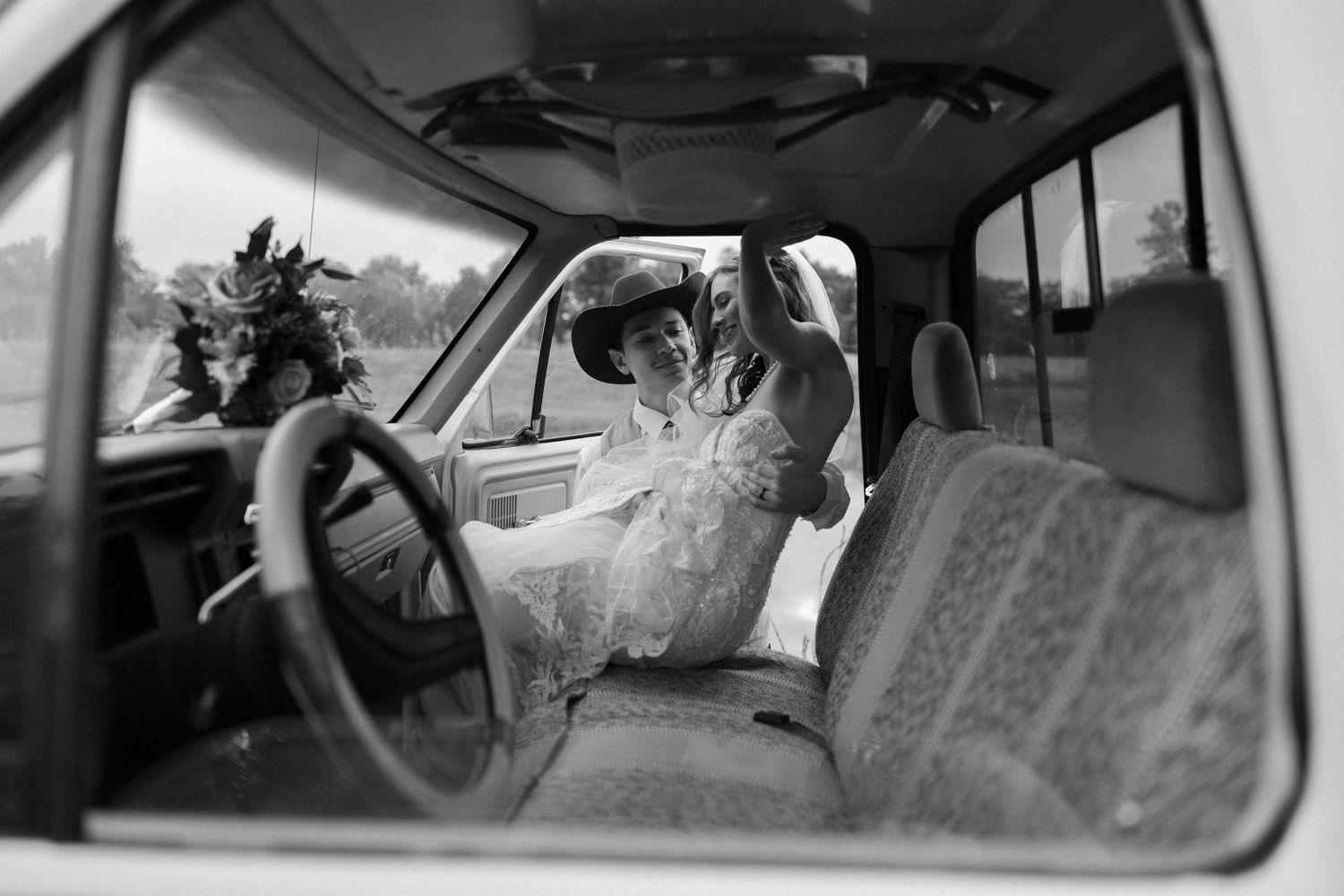 A nostalgic black and white photo taken through a car window showing a bride at the steering wheel.