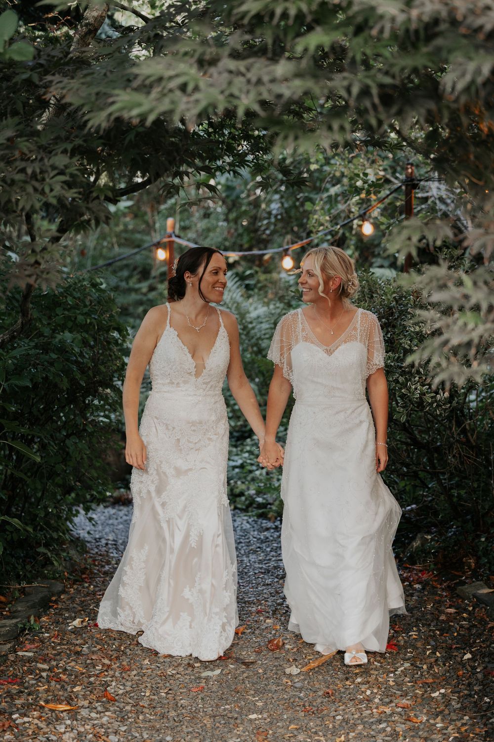 Two brides in white wedding gowns holding hands and walking through a lush garden path together.