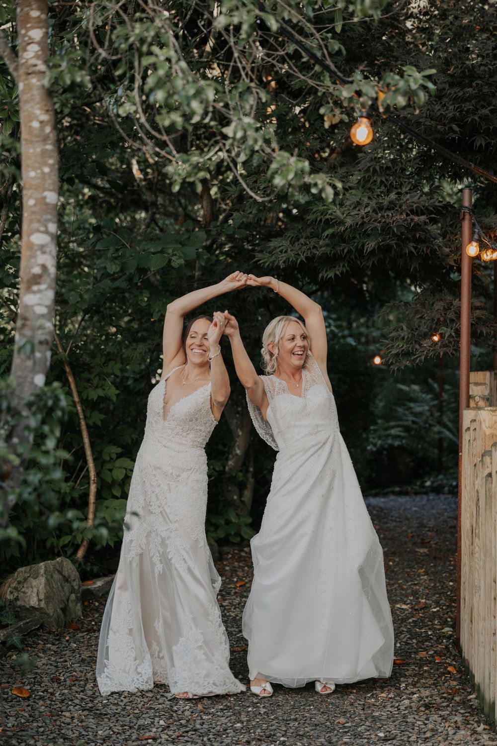 Two joyful brides in white lace gowns dancing with raised arms in a beautiful garden setting.