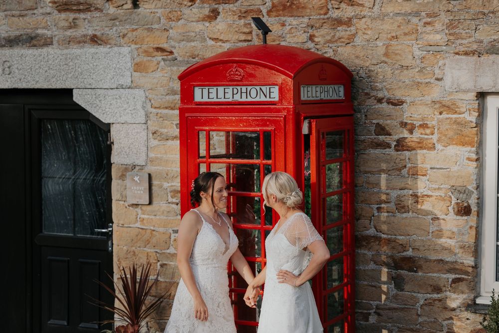 Two brides in elegant white gowns standing together next to a red UK telephone booth outdoors.