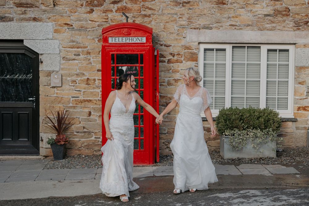 Two brides in white wedding dresses walking hand in hand past a red British telephone box.