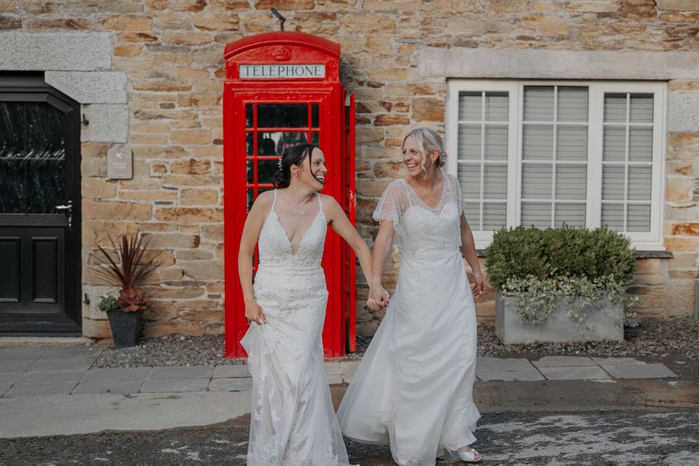 Two brides in white lace wedding dresses hold hands and walk past a red British telephone box.