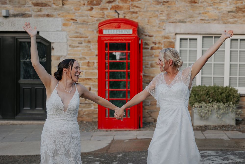 Two brides in white wedding dresses celebrate joyfully with arms raised beside a red telephone box.