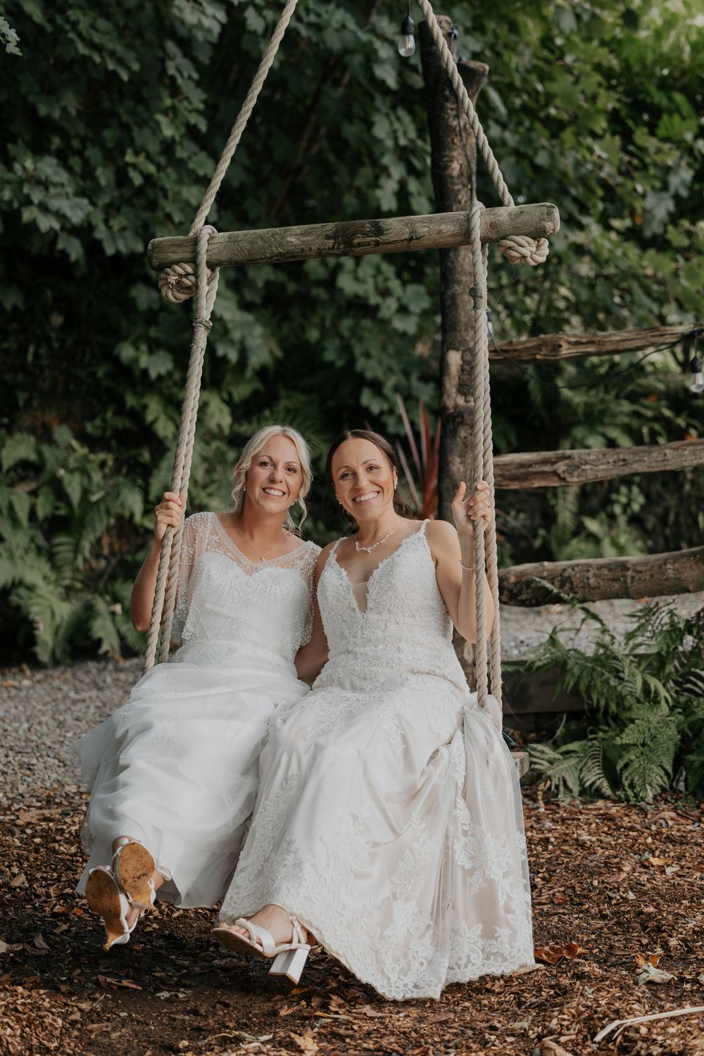 Two brides in white lace wedding dresses smile while sitting together on a rustic rope swing.