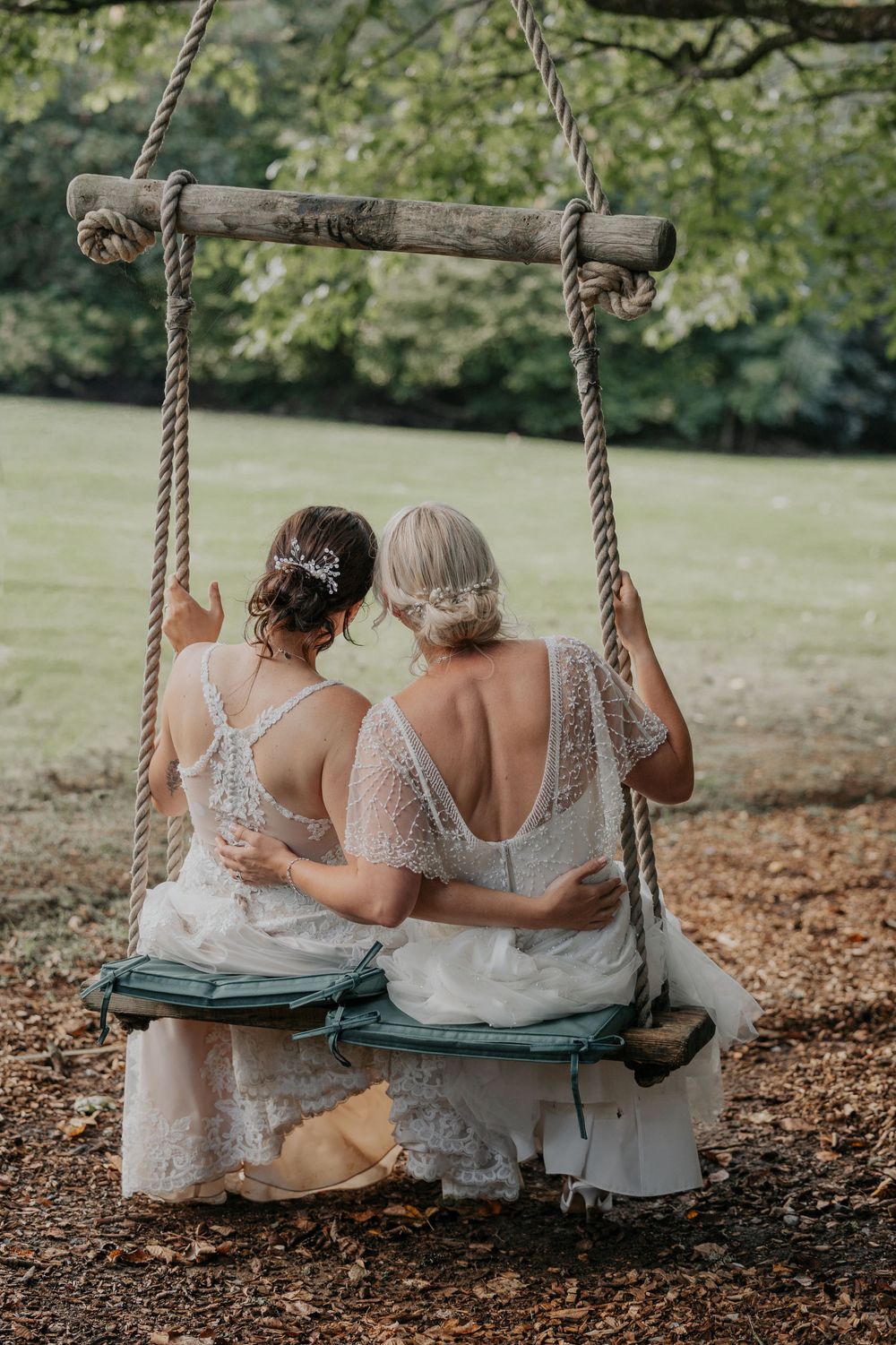 Two brides in backless white lace wedding gowns sit arm in arm on an outdoor swing.