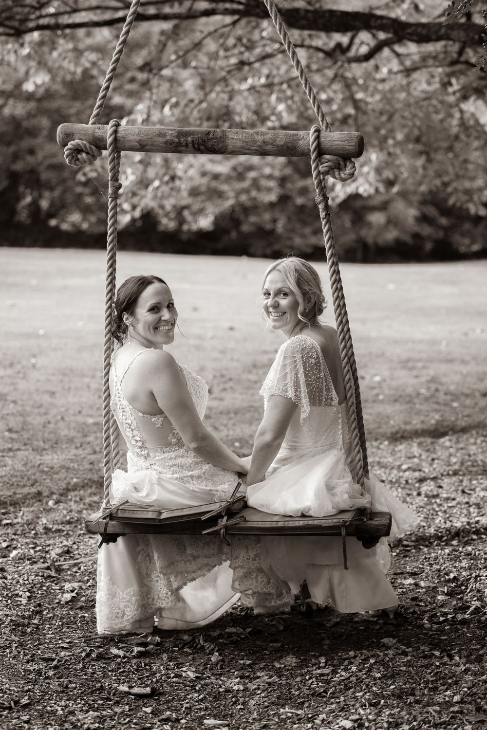 Two brides in white lace gowns sit on a rustic rope swing, holding hands in a romantic black and white wedding photo.