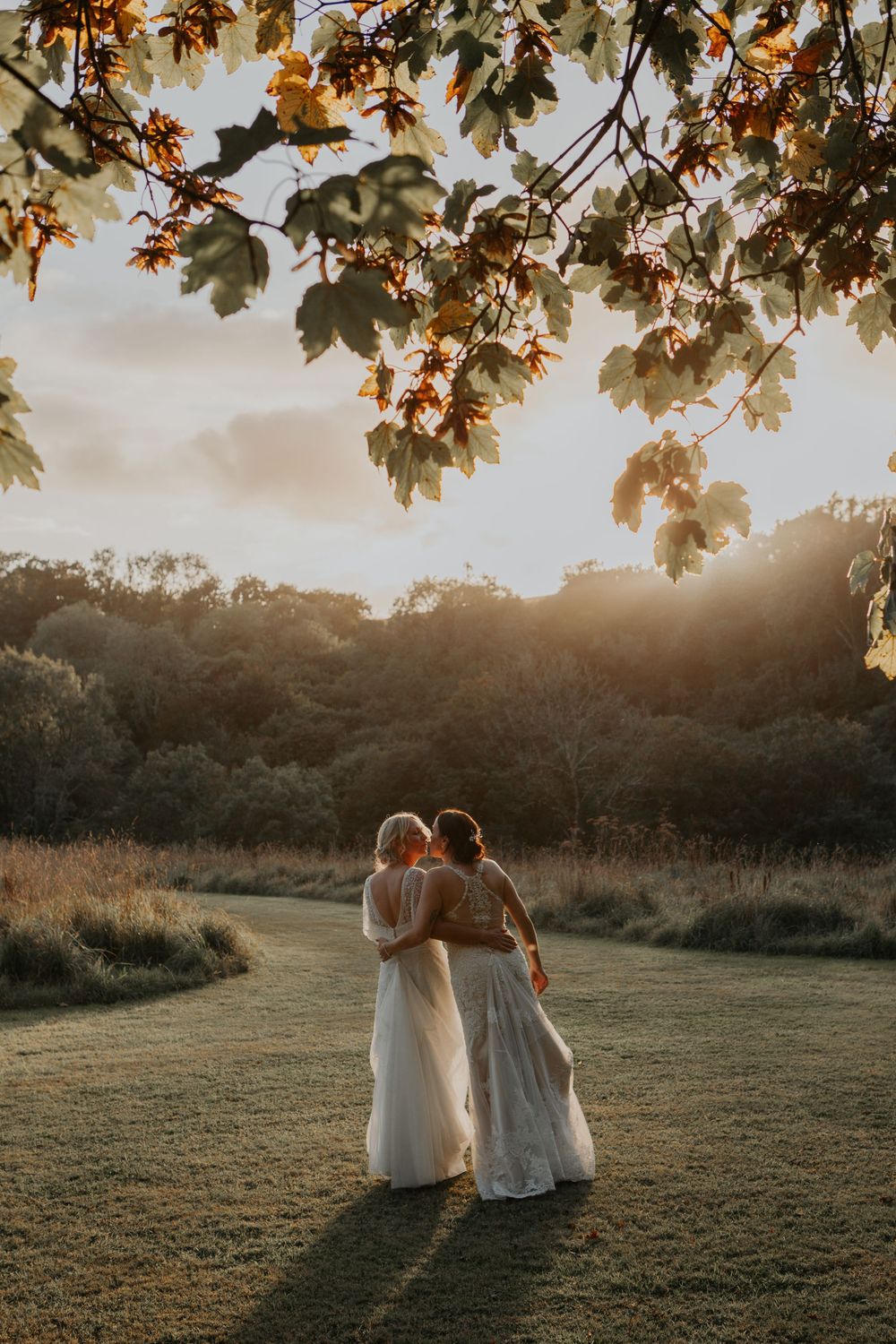 Two brides in white wedding gowns embrace while walking through a sunlit meadow at golden hour sunset.