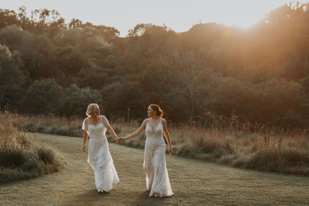 Two brides in white gowns walk hand in hand along a sunlit path surrounded by warm golden hour light.