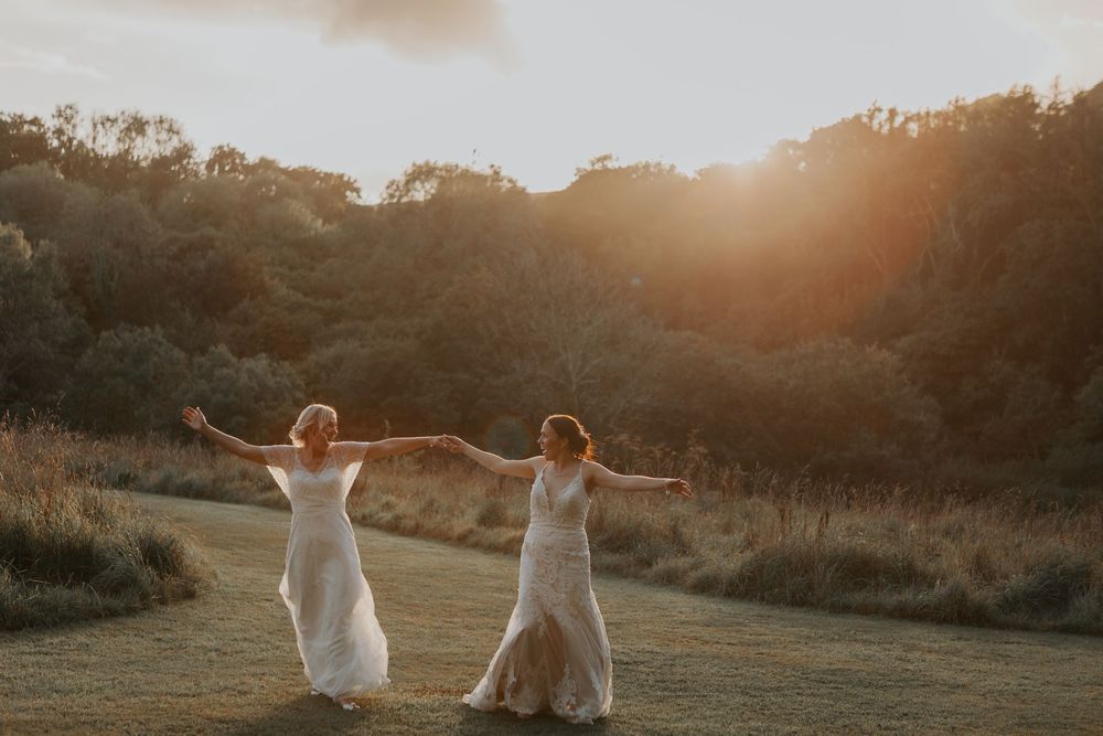 Two brides in white wedding dresses joyfully spin with arms outstretched in a sun-drenched meadow at sunset.