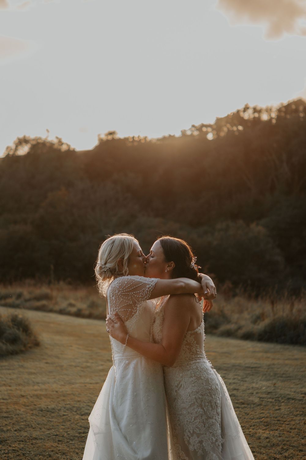 Two brides share a romantic kiss while embracing in a glowing golden sunset meadow on their wedding day.
