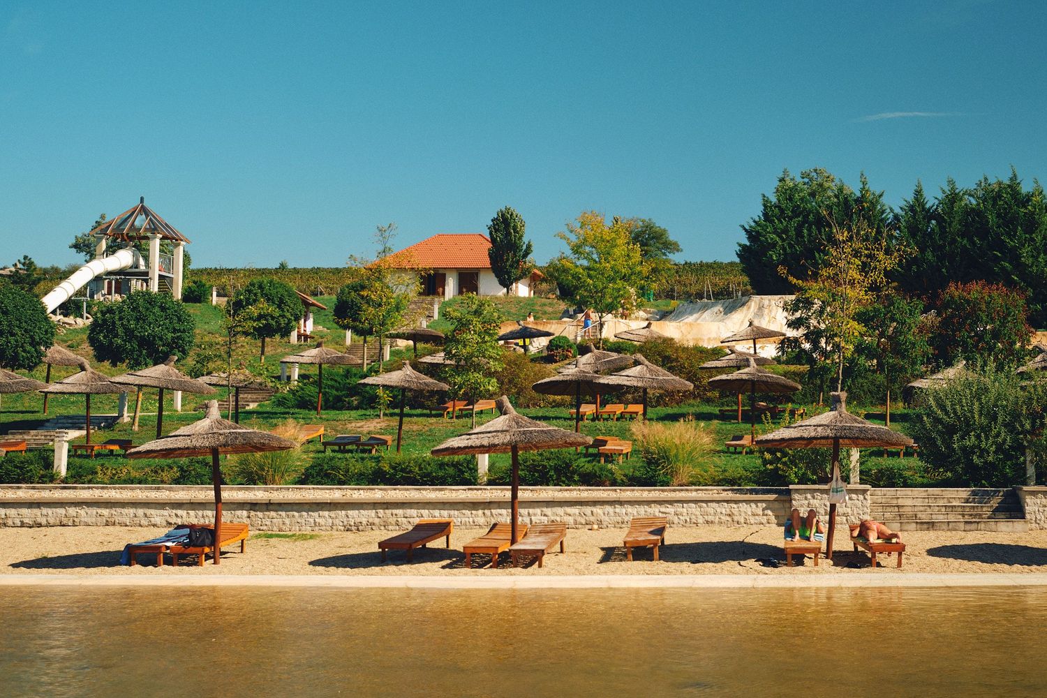 Beach resort with lounge chairs and umbrellas along sandy shore with hillside cottages and trees in background.
