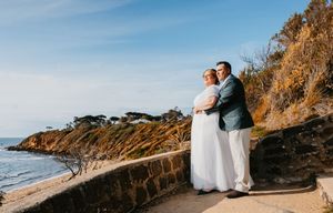 Couple in wedding attire embracing on scenic coastal cliffs with ocean views at sunset.