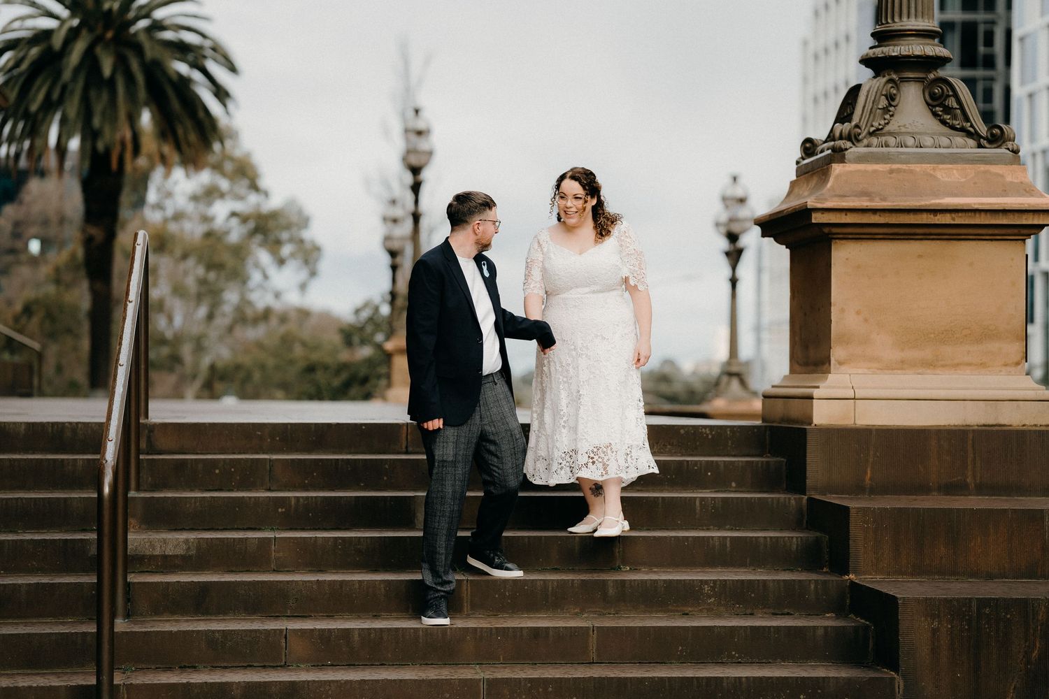 A couple in wedding attire sits together on ornate stone steps in a romantic outdoor setting.