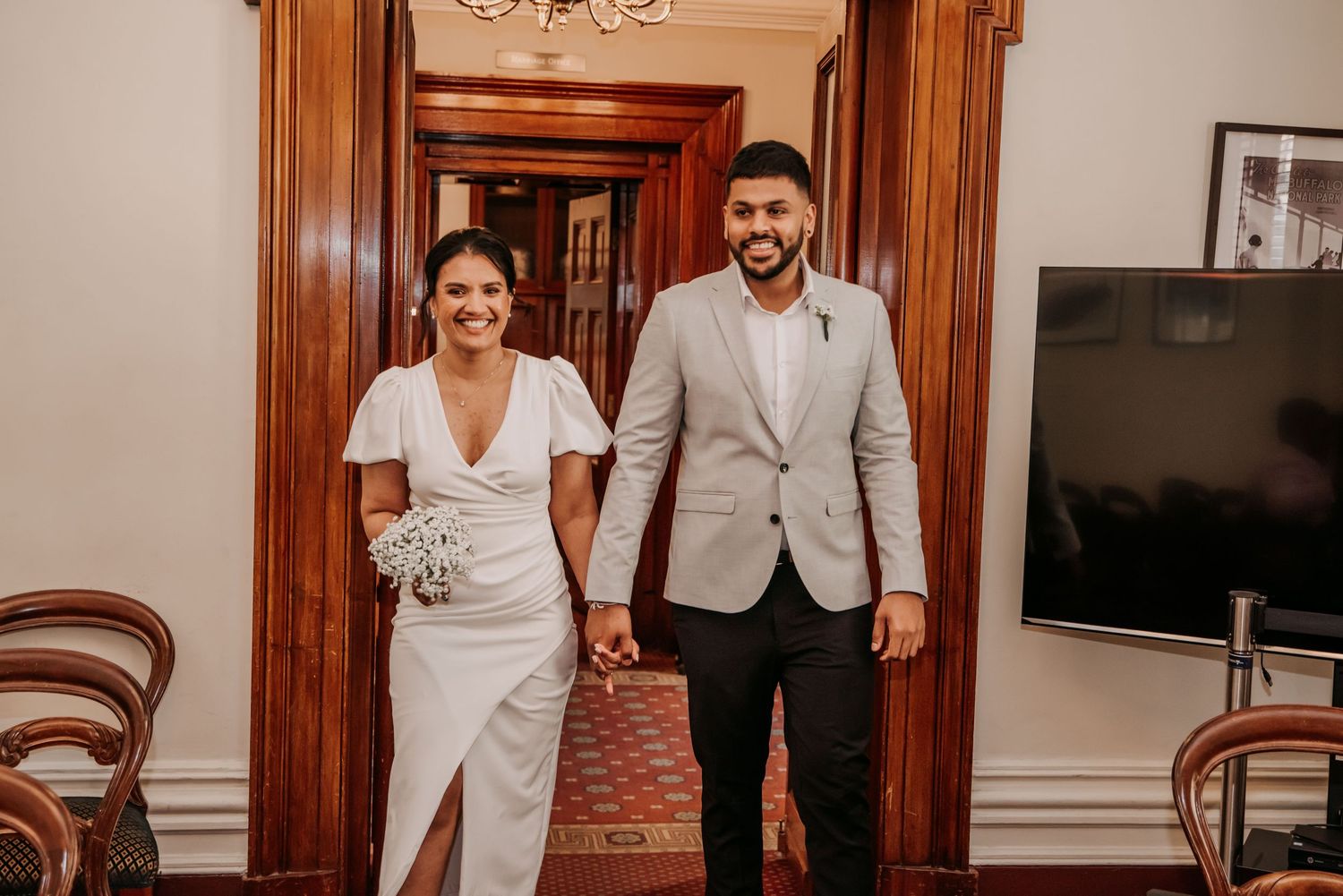 A couple in elegant white and beige formal attire walk hand in hand through an ornate hallway with wood paneling.