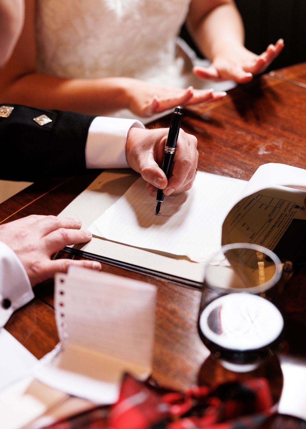 Groom signing wedding certificate with pen at wooden table, bride gesturing nearby, candle and documents visible.