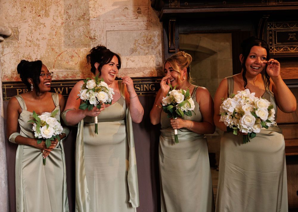 Four bridesmaids in sage green satin gowns laughing and holding white floral bouquets at a wedding ceremony.