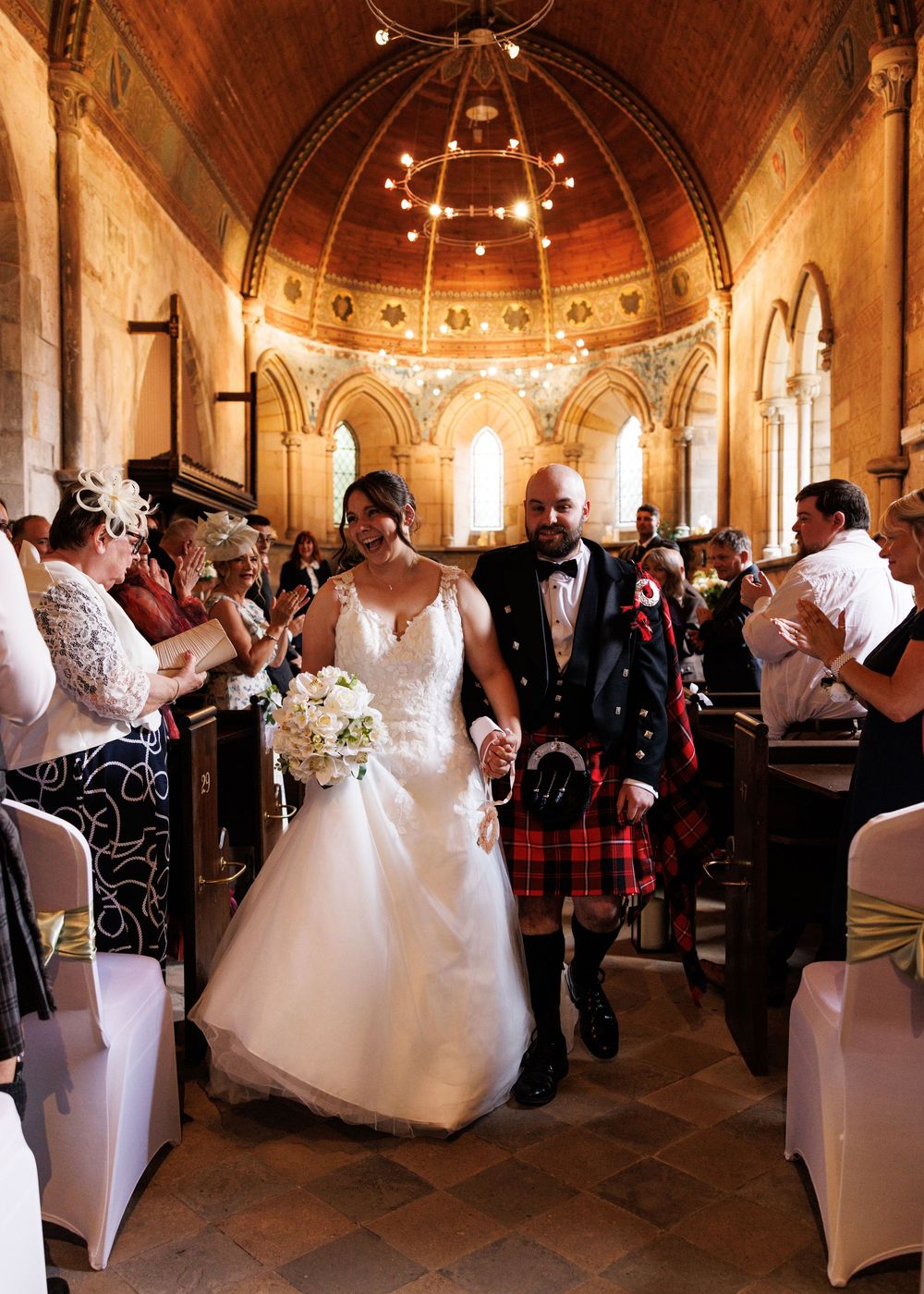 Newlyweds walk down church aisle, groom in red tartan kilt, bride holding white bouquet, guests applauding.