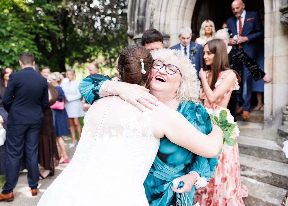 Bride in white lace gown hugging a smiling woman in teal dress outside a church wedding celebration.