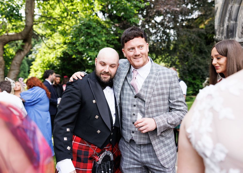 Two men posing at Scottish wedding, one in red tartan kilt and one in grey checked suit, outdoors.